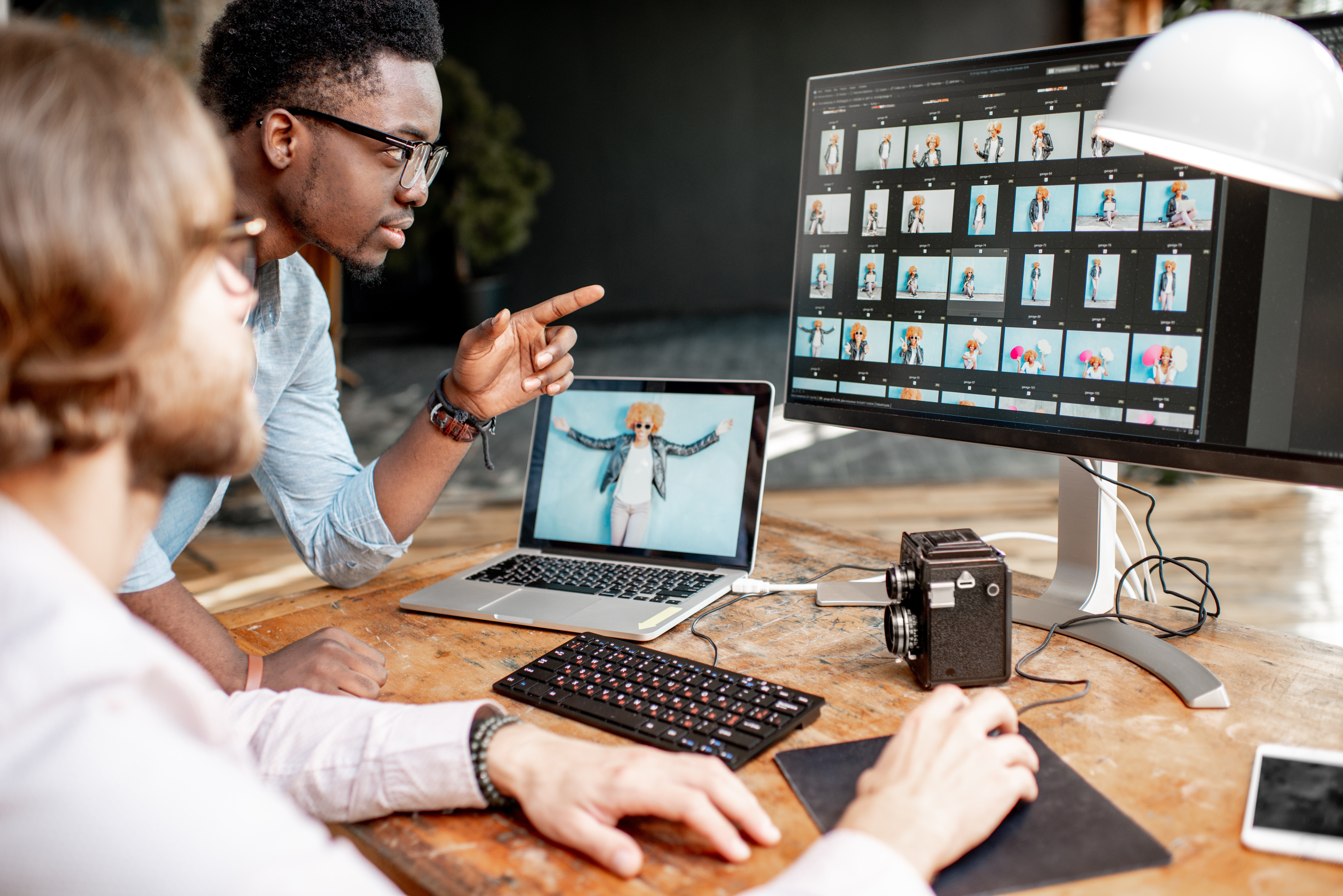 two people pointing at the monitor with a camera and laptop on the table